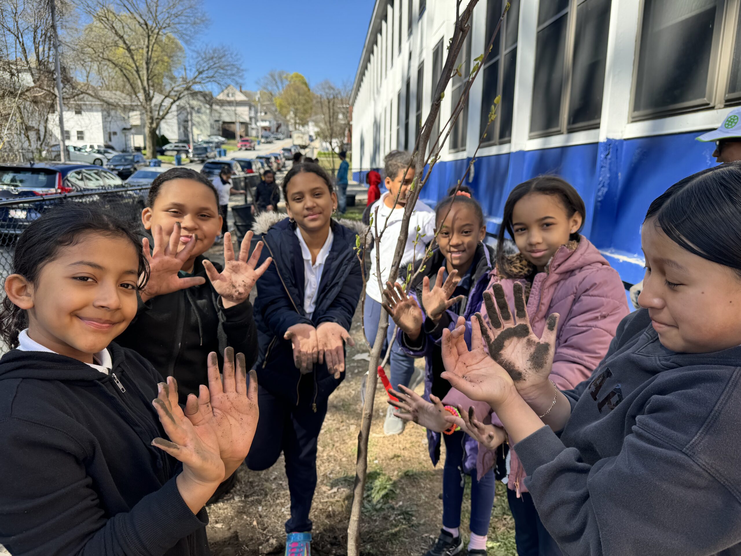 A group of young students wave at the camera as they stand surrounding a newly planted tree.