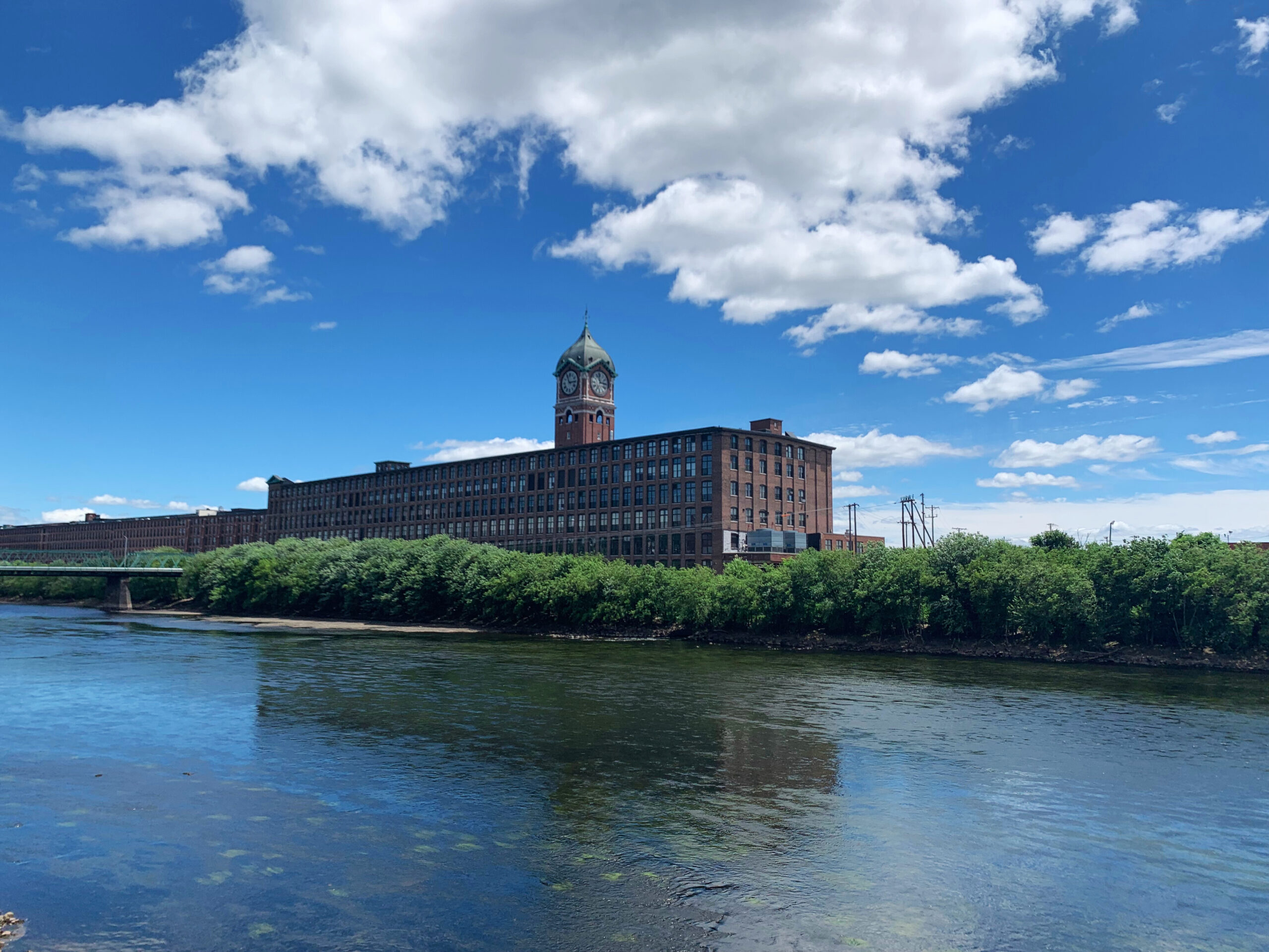 Scenic view of the gently flowing Merrimack River with the historic Ayer Mill Clocktower standing in the background, surrounded by lush greenery.